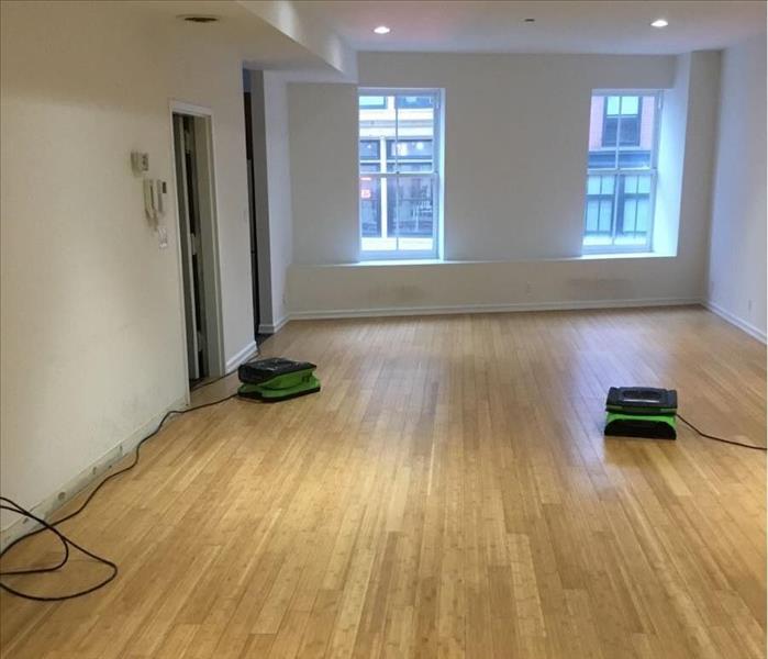 Third-floor living room with wet drywall under windows, wood floors, a removed baseboard, and two air movers operating in it